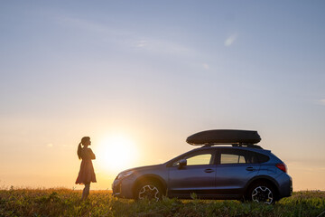 Dark silhouette of lonely woman relaxing near her car on grassy meadow enjoying view of colorful sunrise. Young female driver resting during road trip beside SUV vehicle