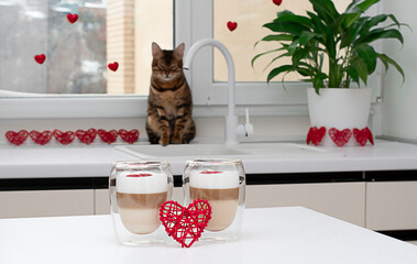 two cups of latte on a table with a red heart against the background of a window and a sleeping bengal cat