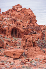 Overton, Nevada, USA - February 24, 2010: Valley of Fire. Closeup portrait of cave-like hole in red rock peak with cracks, black spots and weed crawling up from desert floor. Gray cloudscape.