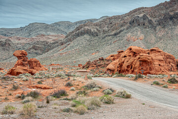 Overton, Nevada, USA - February 24, 2010: Valley of Fire. Gray road meanders between small red rock outcrops with gray mountain range in back under blue cloudscape. Dry desert floor.