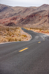 Fototapeta na wymiar Overton, Nevada, USA - February 24, 2010: Valley of Fire. Black asphalt uneven road with yellow lines pulls a line in dry desert landscape with brown mountain range in back under gray sky.