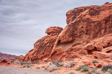 Overton, Nevada, USA - February 24, 2010: Valley of Fire. Red rock cliff with holes, cracks, lines, and black spots on dry desert floor with weeds under thick gray cloudscape.