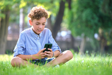 Young boy playing game on his smartphone outdoors in summer park. Addiction from electronic gadgets concept