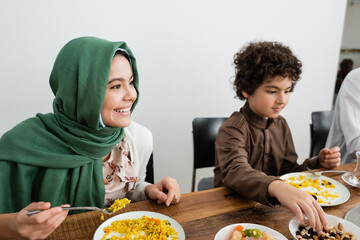 happy muslim girl in hijab eating pilaf near arabian brother.