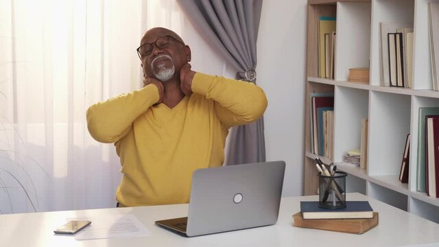 Work break. Tired african man. Warming exercises. Casual senior guy sitting desk with opened laptop stretching neck and hands in light room interior.