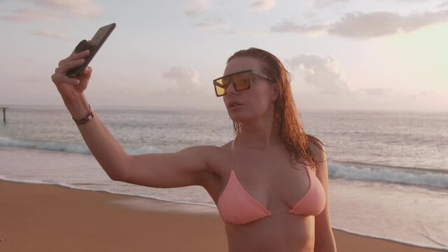 A woman in a bikini posing for selfies with a smartphone on the beach at sunset.
