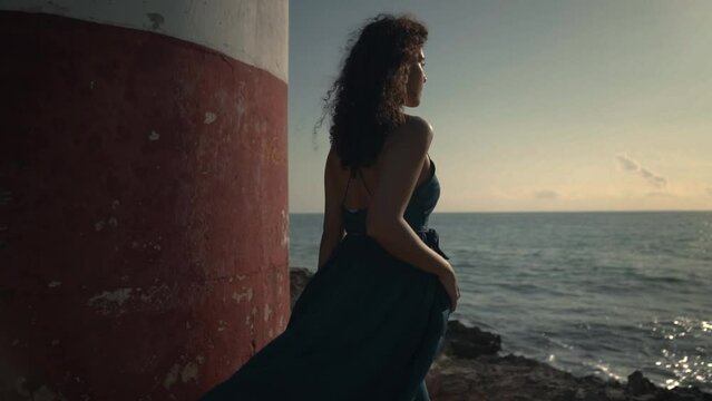 Slow motion m&eacute;dium shot de una mujer joven latina y hermosa con cabello chino casta&ntilde;o, en un vestido azul, caminando sobre las rocas en la playa, a un lado de un faro, mirando el amanecer