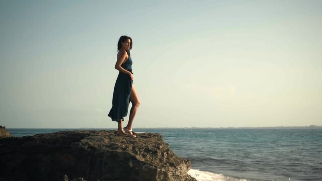 Vista lateral de una mujer latina hermosa de cabello chino con un vestido, parada a la orilla de las rocas en la playa, disfrutando de la vista y el viento  al amanecer