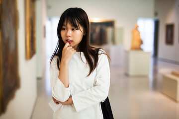 Thoughtful chinese female visitor with guide-book looking at artwork painting in modern museum...