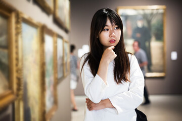 Fototapeta premium Portrait of thoughtful chinese young woman looking at paintings in the museum