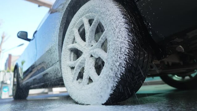 Close-up car wheel covering with white foam. Cleaning of automobile tire at car wash service outdoors. Vehicle maintenance and purity concept