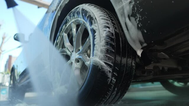 Close-up washing off white foam from car wheel with water. Cleaning of black luxurious automobile outdoors with high pressure washer at car wash service