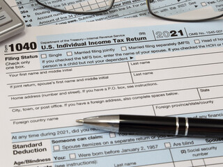 An IRS 1040 tax year 2021 form is shown in 2022, along with an ink pen, calculator, and glasses. The Internal Revenue Service tax filing deadline in 2022 is scheduled for April 18.