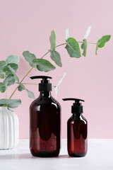 Dark amber glass cosmetic dispenser bottles and eucalyptus branch on pink background. Organic shampoo and shower gel packaging design, branding.