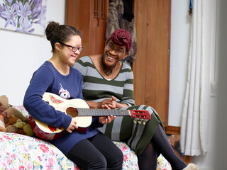 Mother assisting daughter with Down Syndrome playing guitar in bedroom