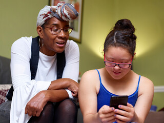 Mother with daughter with Down Syndrome looking at smart phone at home - obrazy, fototapety, plakaty