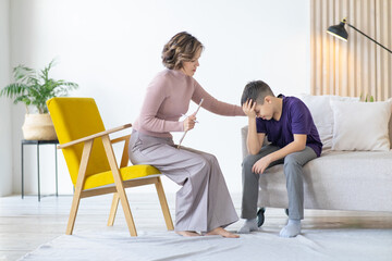 Counseling adolescent psychologist calms and holds adolescent by shoulder. Boy in purple t-shirt is worried and holds his head with his hand, while sitting on couch in minimalistic room.