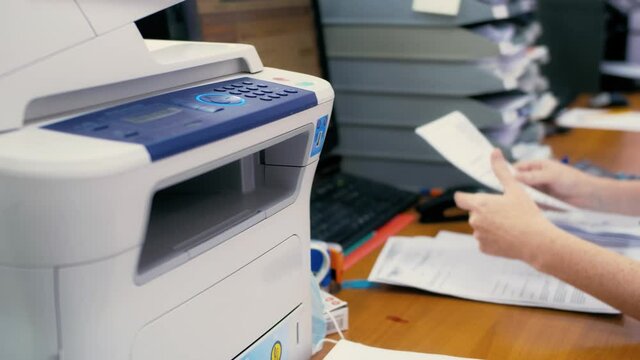 Employee using printer or copier in office. Hand takes printed sheet and continues to work on the computer.