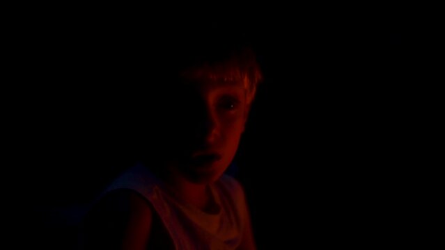 Spooky portrait of a little boy's face lit up by red firelight in the dark outdoors.