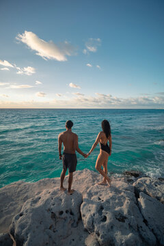 Pareja de j&oacute;venes latinos enamorados tomados de las manos en la playa, sobre las rocas, disfrutando de la vista