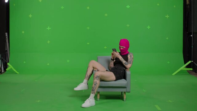 Young woman in pink balaclava with smartphone in hands. Hooligan girl in mask sitting in chair and using mobile phone on green screen background . Chroma key . 4k uhd video