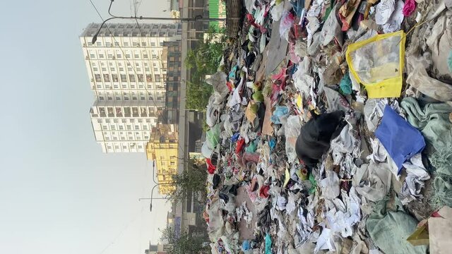 Vertical view of a garbage dump with a black dog sitting on the trash and eating stale food of the dump with the view of a multi storied building  in the background in Dhaka, Bangladesh.