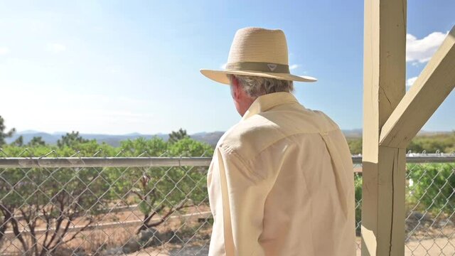 Old Man looks into distance from his patio towards orchards and mountains, then turns around and walks away, camera following.