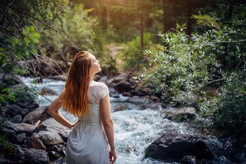 Fototapeta na wymiar Portrait long-haired brunette in white dress posing against small mountain river and green trees. Beautiful young woman on the forest stream shore.