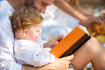 Happy Caucasian family couple with little child boy enjoy outdoor lifestyle activity on summer travel vacation. Father reading a book with little baby son while picnic together on tropical beach.