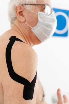 Older latin and hispanic male with a kinesiology tape on his shoulder and wearing a mask attending therapy. Vertical photo.