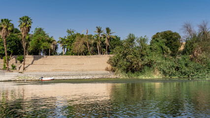 The bank of the Nile is fortified with a stone wall. The boat is standing on calm water. Lush green vegetation against a blue sky. Reflection. Egypt