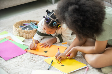Happy African cute child girl kid sibling lying on the floor using color pencil drawing and painting on the paper together. Little sister having fun indoor lifestyle leisure activity together at home.