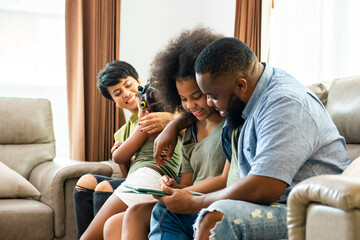 Smiling African parents and two little daughter sitting on sofa in living room using digital tablet play games or watch movie together. Happy family enjoy weekend activity with technology at home