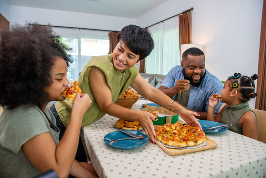 African family parents and two little daughter eating fried chicken and pizza on table for dinner together. Father and mother and cute child girl kid enjoy eating and sharing a meal together at home