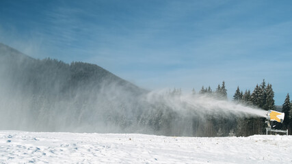 Snow cannon makes artificial snow on bright sunny day at ski resort. On back winter forest, blue sky. Snowmaking systems sprays water to produce snow. Preparation of ski track for skiing, winter sport