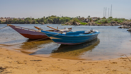 Naklejka na ściany i meble Two bright wooden boats are tied up near the bank of the Nile. There is yellow sand on the beach. Shadows and reflections on clear water. Green vegetation, boulders against the blue sky. Egypt. Aswan