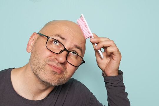 Bald, bearded man with glasses is scratching his head with a child's comb on a monochrome background. A man with green eyes in a black T-shirt with a long sleeve combs his bald head.