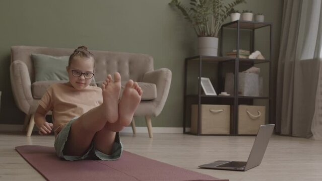 Slowmo shot of cute 11-year-old girl in eyeglasses practicing yoga on mat at home using laptop with online lesson
