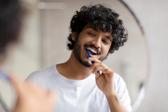 Oral care concept. Young indian man cleaning teeth with toothbrush, smiling to his reflection in mirror