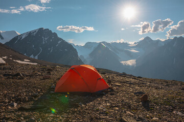 Awesome alpine landscape with orange tent with view to high snow mountains and large glacier. Bright sun in cloudy sky above snowy mountain range. Tent with top view to glacier tongue shining in sun.