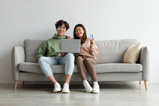 Online shopping. Asian couple sitting on couch at home, using laptop and credit card, purchasing goods in web store
