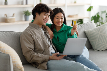 Positive Asian family doing online shopping with credit card and laptop, sitting on sofa and cuddling at home