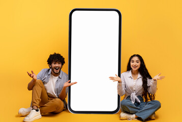 Surprised indian couple sitting near huge smartphone with blank screen over yellow studio...