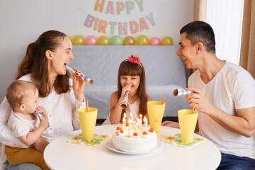 Obraz na płótnie Canvas Indoor shot of happy little girl celebrating her birthday with family, with cake and candles, child receiving congratulations at home, blowing party horns together with parents.