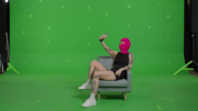 Young woman in pink balaclava with smartphone in hands. Hooligan girl in mask sitting in chair and taking selfie self portrait photo on green screen background . Chroma key . 4k uhd video