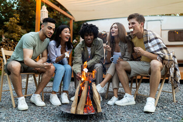 Diverse millennials having outdoor picnic near trailer, frying marshmallows on fire in evening,...