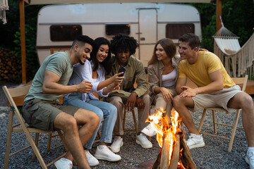 Diverse friends sitting near campfire in evening, having autumn camping trip with RV, watching video online on cellphone