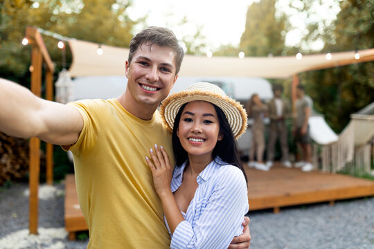 Young happy diverse couple taking selfie at camp, resting with friends near trailer, spending autumn weekend outdoors - Powered by Adobe