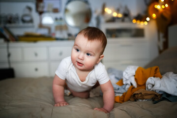 Fototapeta premium funny european chubby baby crawling among clothes on bed, children's clothes and conscious consumption, lot of clothes and child, big pile of clean clothes and baby playing, childhood and emotions