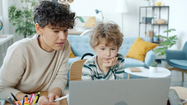 Young woman explaining science to smart son looking at laptop screen and talking in apartment. Modern technology and homeschooling concept.